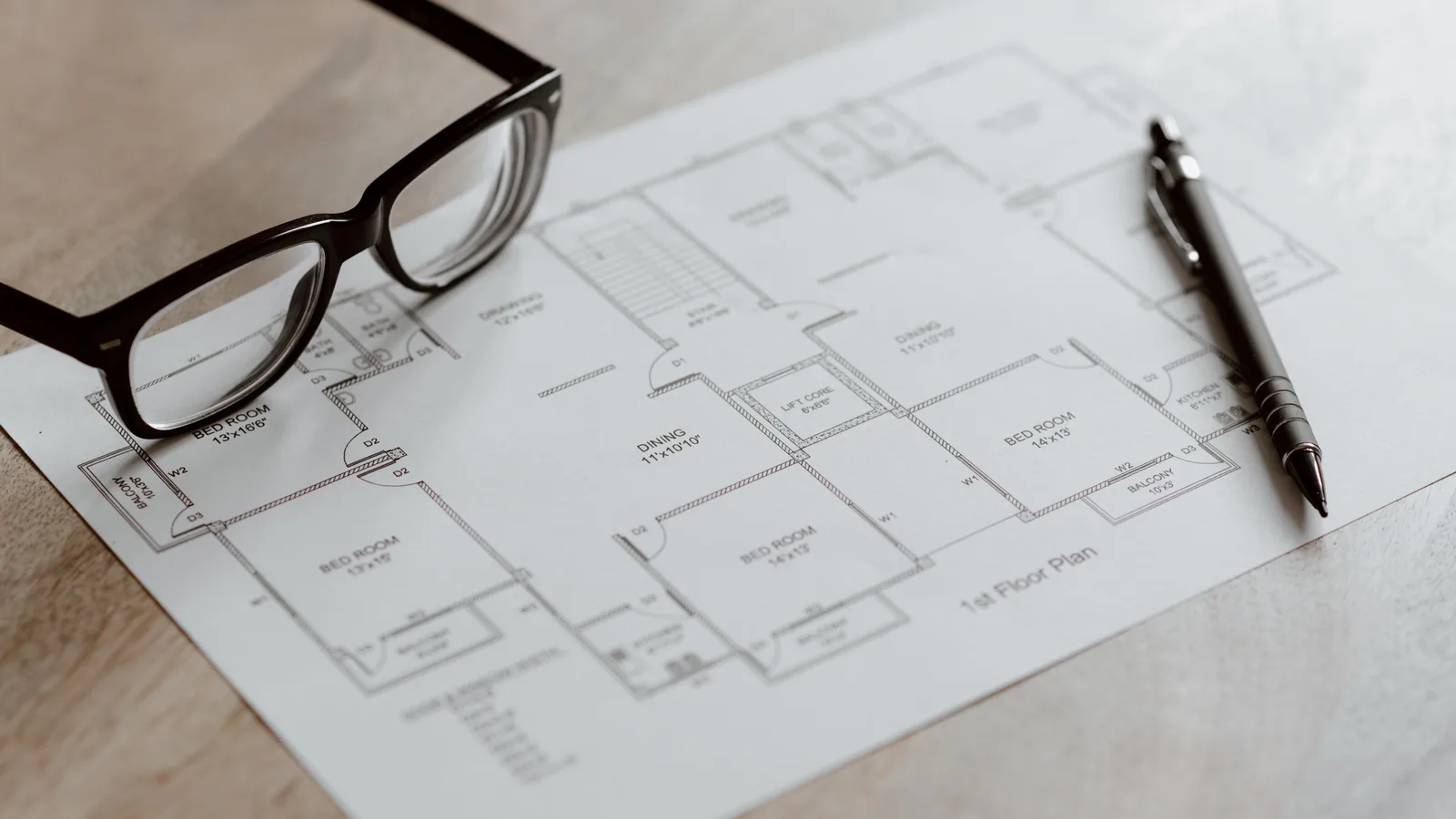 Composition of plan of room and black pen with eyeglasses placed on wooden table at daytime