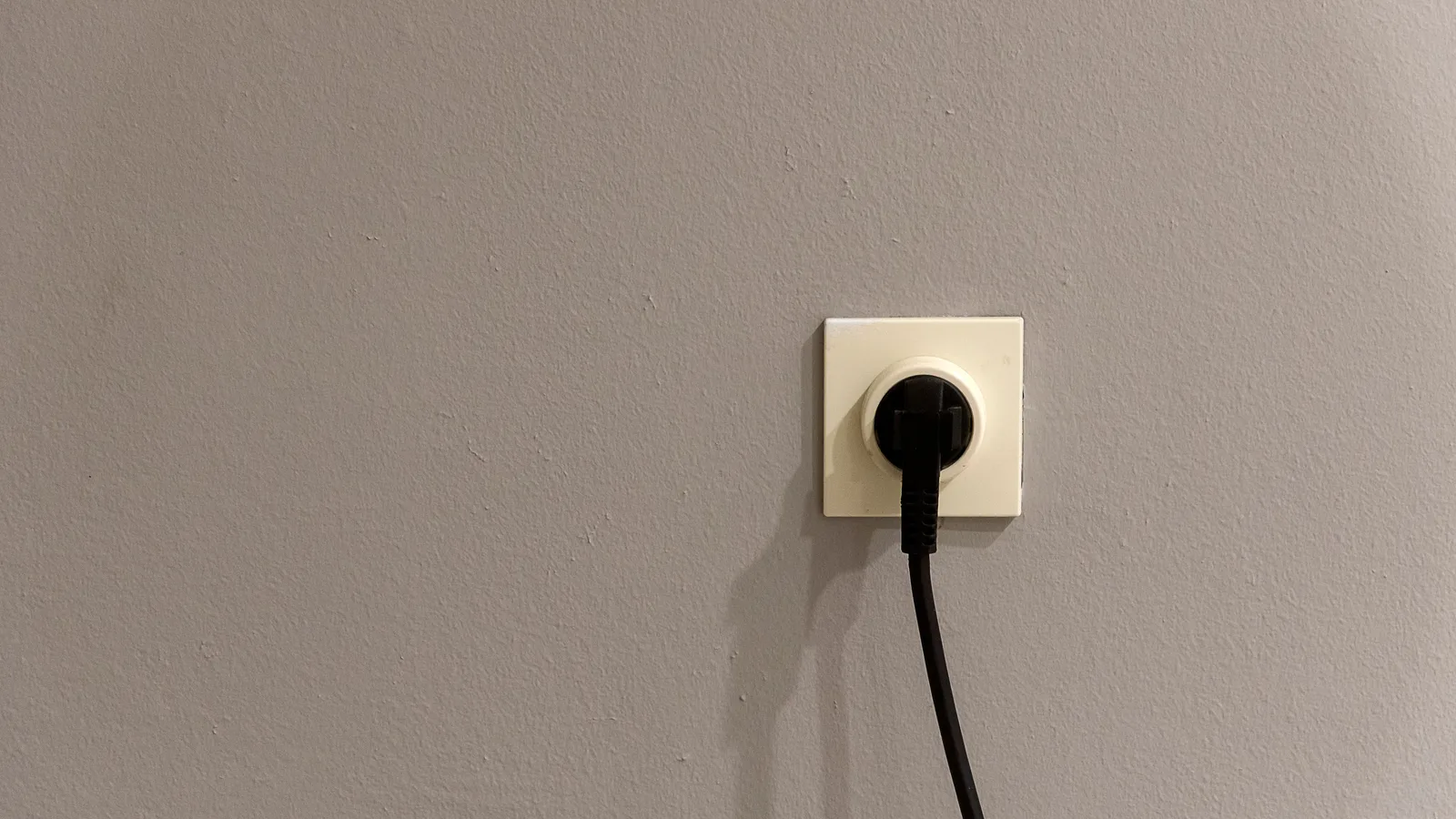 A black plug inserted into a white wall socket on a plain grey wall.