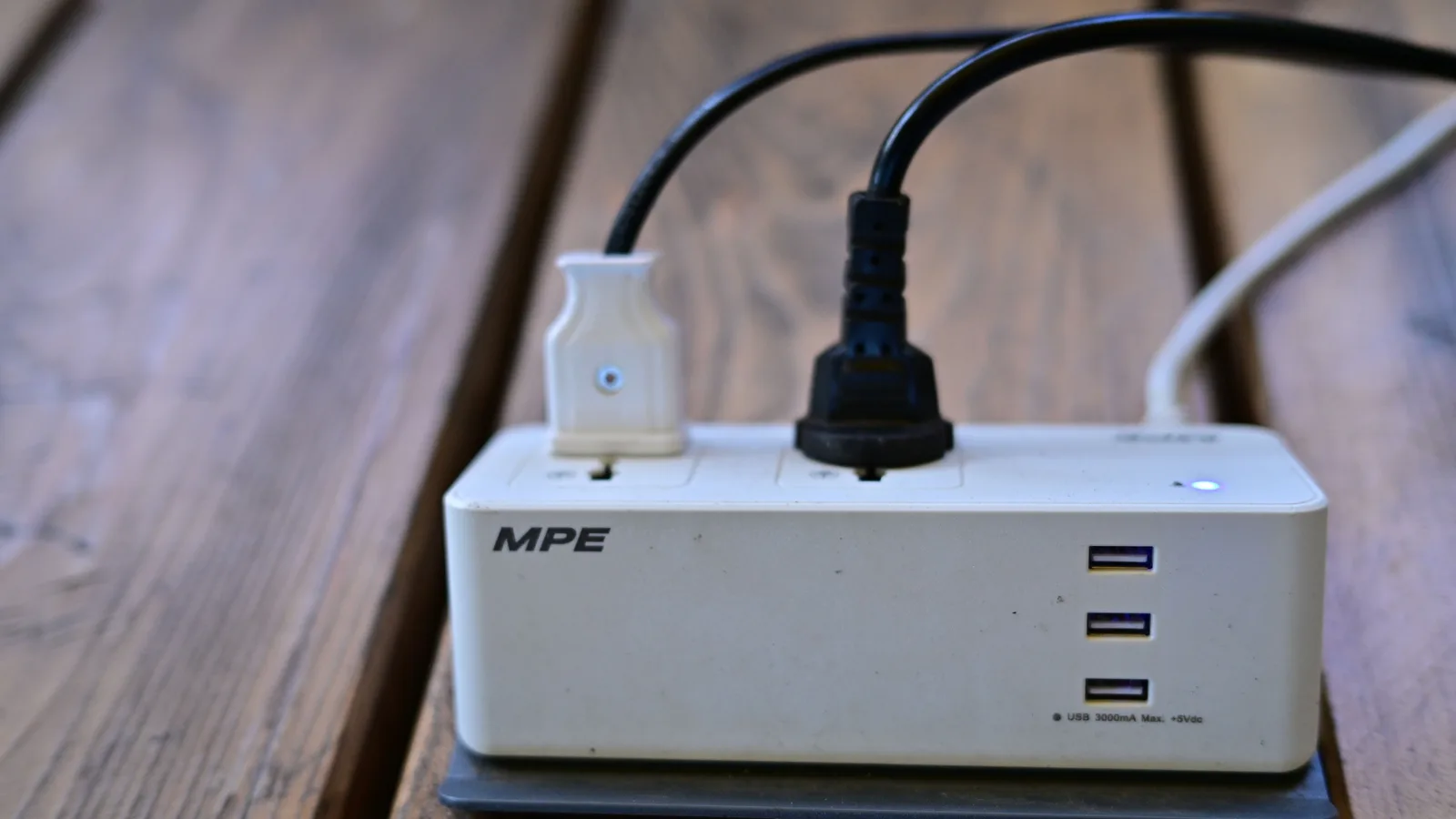 Close-up of a white MPE USB power/charging strip with two plugged-in cables and three blue USB ports on wooden slats